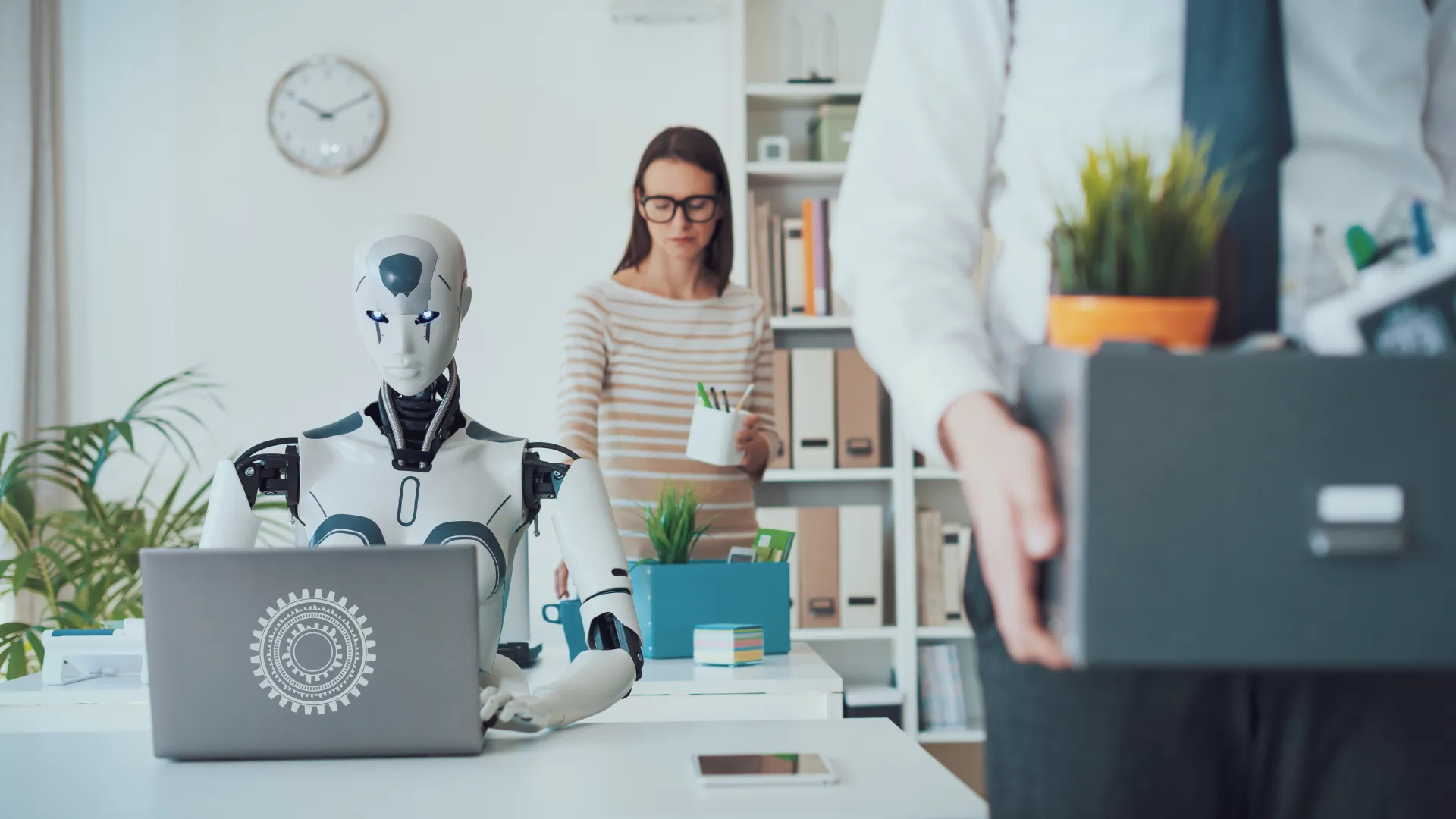 Ein humanoider Roboter arbeitet im Büro am Laptop, während zwei Beschäftigte ihre Sachen in Kisten packen und gehen.